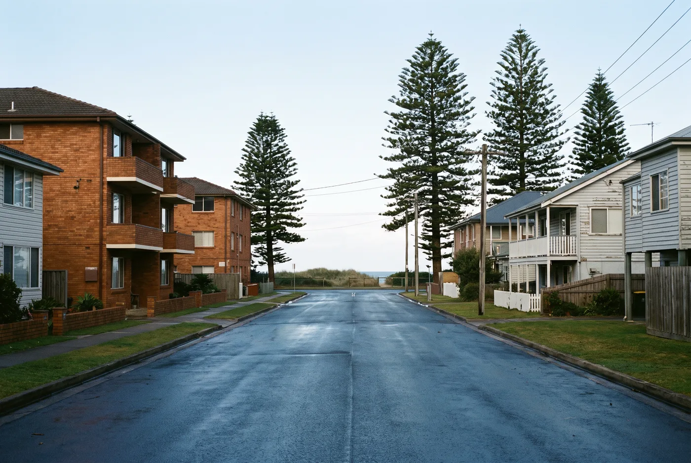 A flat Narrabeen street with three-storey brick walk-up flats on one side, weatherboard houses on the other, Norfolk Island pines, and the sea visible at the end of the street